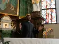 Duchowny w czerni, głoszący kazanie. W tle ołtarz, witraż i obrazy. To jego kapłaństwo.