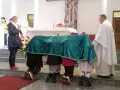 Dzieci klęczą pod zielonym welonem, odmawiając modlitwę do Matki Bożej z Guadalupe. Kapłan czuwa obok.
