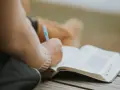 Osoba pisze w otwartej księdze, być może dziennik. W tle widać fragmenty ubrania. To moment refleksji, gdzie żal doskonały może znaleźć swoje ujście w słowach.
