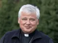 Uśmiechnięty jałmużnik papieski w sutannie i czarnym polarze, na tle zieleni.