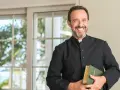 Uśmiechnięty mężczyzna w sutannie, trzymający Biblię. Wygląda jak prefekt, gotowy do wygłoszenia kazania.