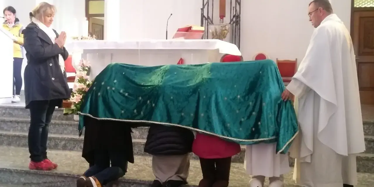 Dzieci klęczą pod zielonym welonem, odmawiając modlitwę do Matki Bożej z Guadalupe. Kapłan czuwa obok.