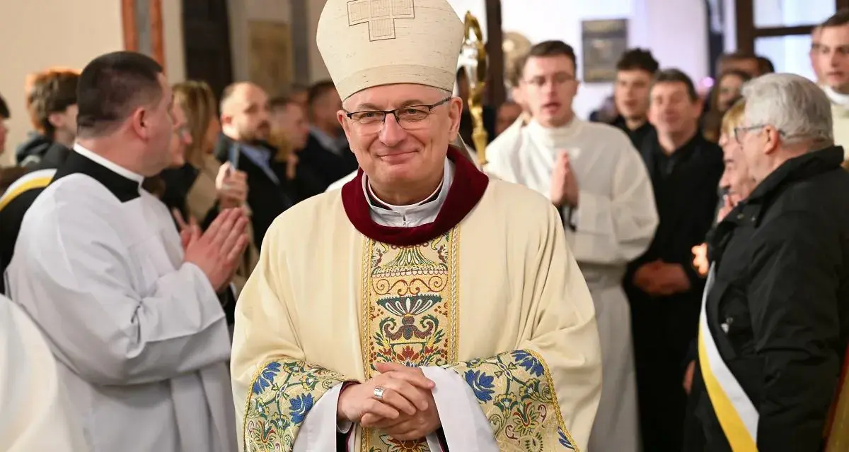 Uśmiechnięty biskup w stroju pontyfikalnym, otoczony przez wiernych i duchownych. W tle widać fragmenty kościoła, a na pierwszym planie szczegóły szat.