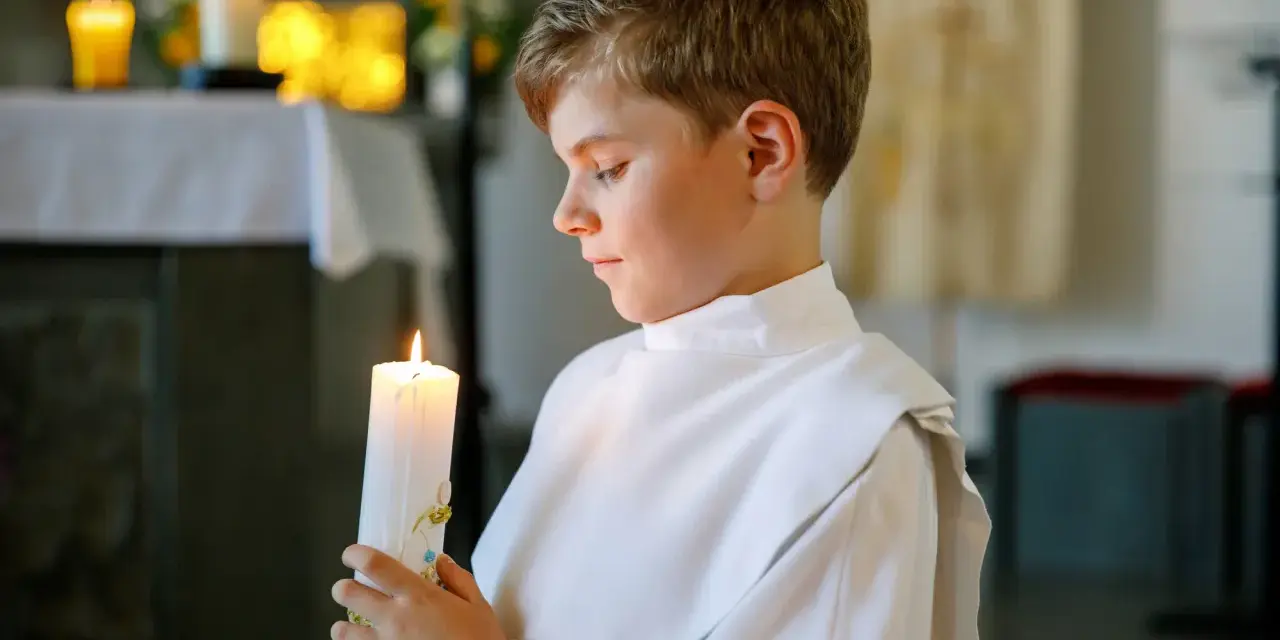 Chłopiec w albie trzyma zapaloną świecę podczas swojej pierwszej komunii świętej.