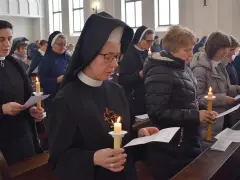 Siostra zakonna z zapaloną świecą i modlitewnikiem w kościele. Wierni w ławkach trzymają świece i kartki.
