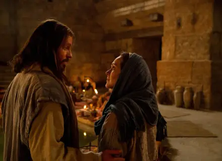 Scena z Kany Galilejskiej: Jezus rozmawia z kobietą, w tle widać gości weselnych i świece.