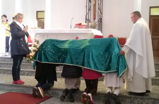 Dzieci klęczą pod zielonym welonem, odmawiając modlitwę do Matki Bożej z Guadalupe. Kapłan czuwa obok.