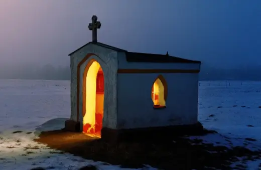 Kapliczka z krzyżem na dachu, oświetlona od środka, w zimowym krajobrazie. Symbol nadziei i religii.