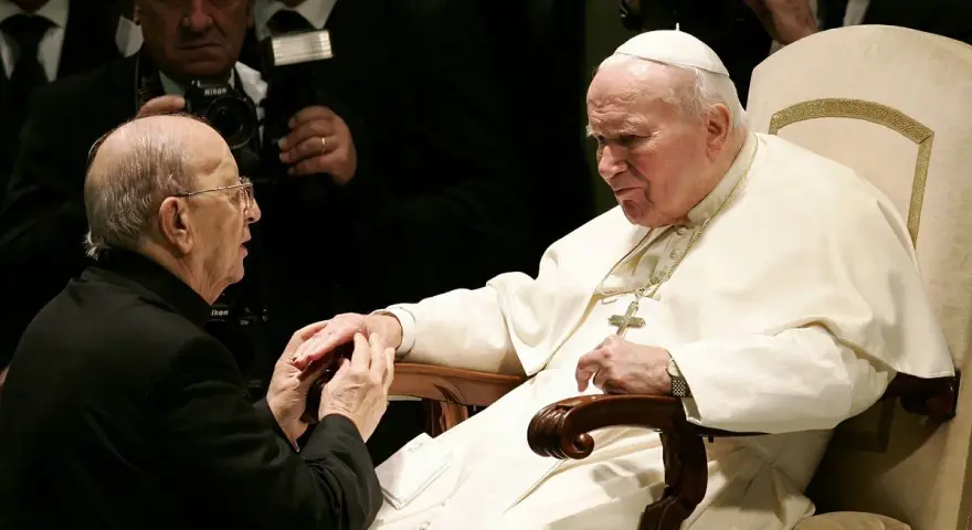 Papież Jan Paweł II w białej szacie, klęczący mężczyzna w czerni. W tle widać fotografów. To obraz duchowej siły, jakiej doświadczają legiony Chrystusa.