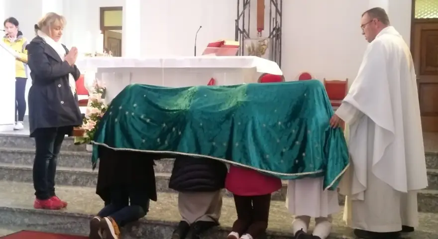 Dzieci klęczą pod zielonym welonem, odmawiając modlitwę do Matki Bożej z Guadalupe. Kapłan czuwa obok.