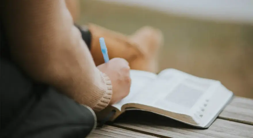Osoba pisze w otwartej księdze, być może dziennik. W tle widać fragmenty ubrania. To moment refleksji, gdzie żal doskonały może znaleźć swoje ujście w słowach.