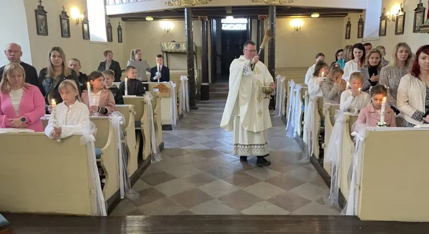 Uroczyste odnowienie przyrzeczeń chrzcielnych w kościele. Dzieci z zapalonymi świecami siedzą w ławkach, a ksiądz błogosławi zgromadzonych.