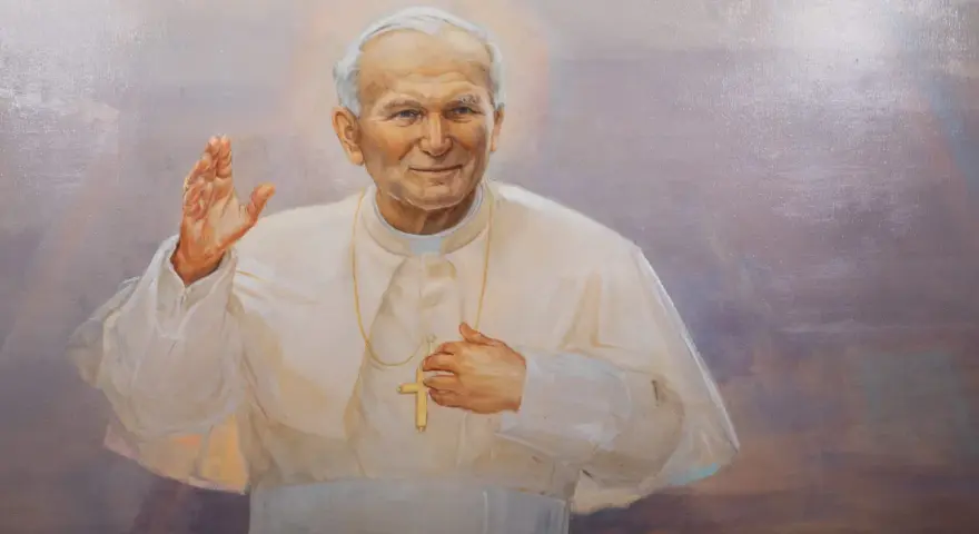 Obraz przedstawia Jana Pawła II w białej szacie, z podniesioną prawą dłonią i krzyżem na piersi. Wierni odmawiają modlitwę do Jana Pawła II o uzdrowienie.