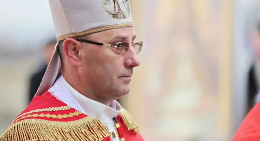 Biskup w stroju pontyfikalnym, z mitrą na głowie, w okularach. W tle widać fragment obrazu.