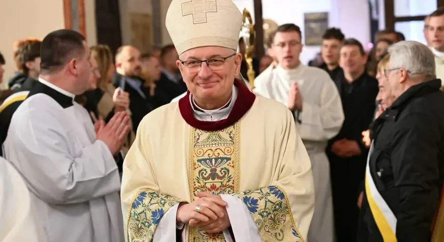 Uśmiechnięty biskup w stroju pontyfikalnym, otoczony przez wiernych i duchownych. W tle widać fragmenty kościoła, a na pierwszym planie szczegóły szat.