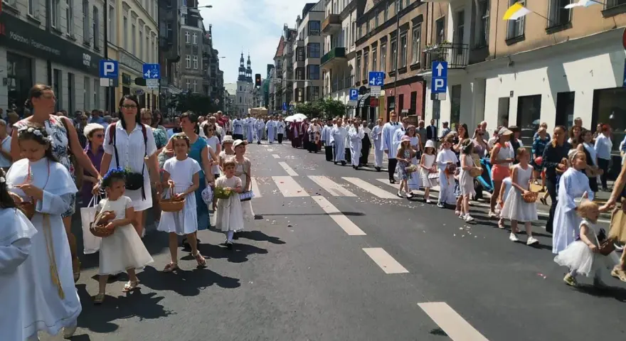 Procesja dzieci w białych strojach, niosących koszyki, idzie ulicą. W tle widać budynki i ludzi, co może symbolizować sekularyzację społeczeństwa.