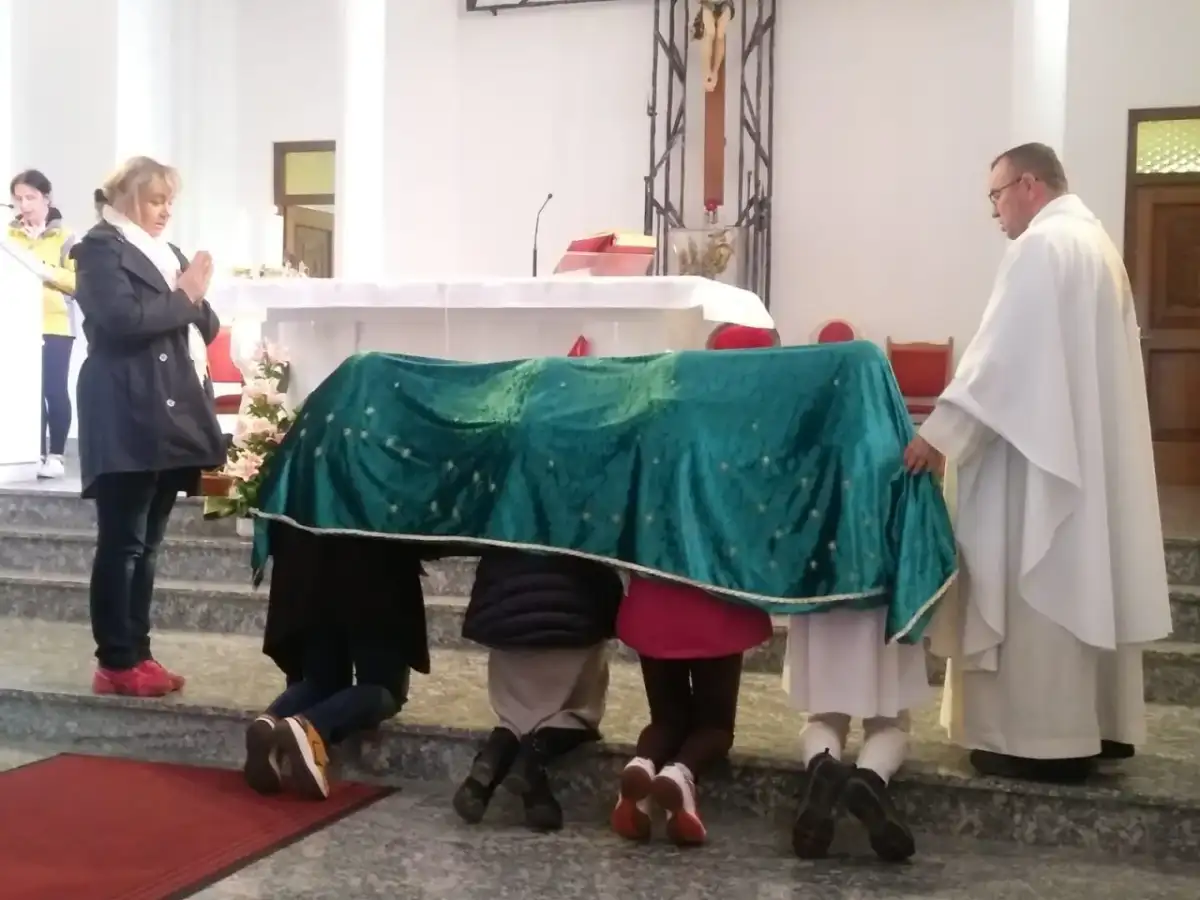 Dzieci klęczą pod zielonym welonem, odmawiając modlitwę do Matki Bożej z Guadalupe. Kapłan czuwa obok.