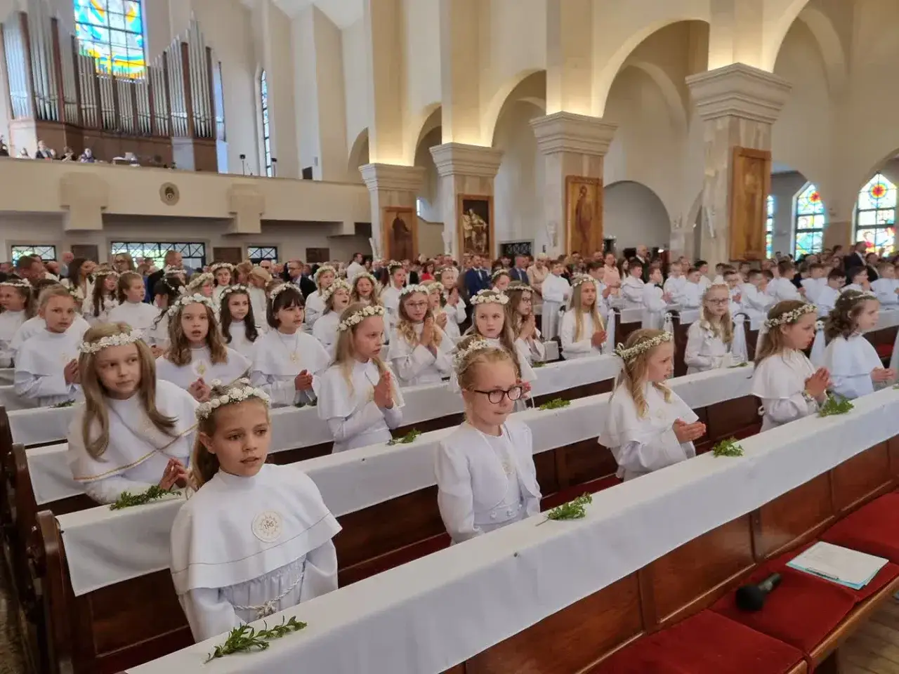 Dzieci w białych strojach, z wiankami na głowach, siedzą w ławkach kościoła podczas uroczystości pierwszej komunii świętej.