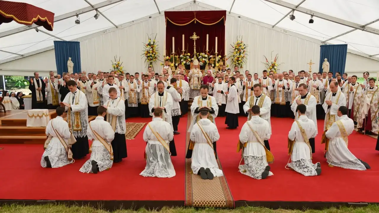 Uroczystość święceń kapłańskich pod namiotem. Klęczący kandydaci otrzymują błogosławieństwo od biskupów i kapłanów.