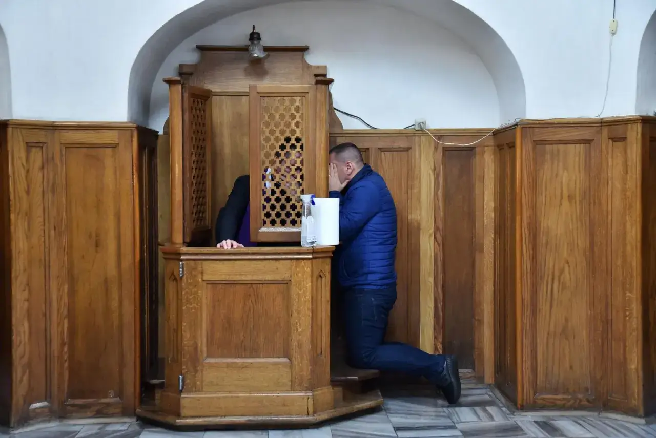 Mężczyzna klęczy przed kratką konfesjonału, zasłaniając twarz. Spowiednik słucha jego żalu.
